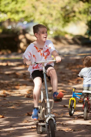 A girl with a blood-stained shirt rides a bike while a smaller child follows on a trike.