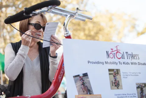 A woman applies her own face paint beside a NorCal Trykers banner about providing mobility to kids with disabilities.