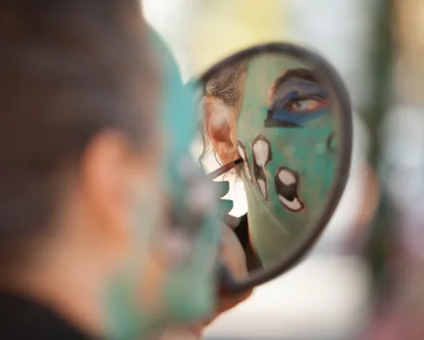 A mirror reflects green face paint mid-application behind a blurred foreground head.