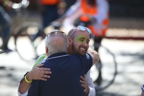 A smiling man with green face paint hugs an older man warmly on the parade route.
