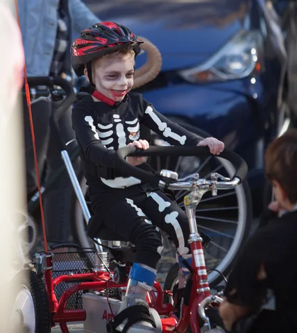 A grinning boy in a skeleton costume and zombie makeup rides an adaptive AmTryke tricycle.