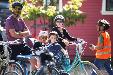 A family in zombie face paint sits on bikes together in front of a red building.