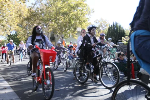 Costumed riders on bikes pedal past a red building.