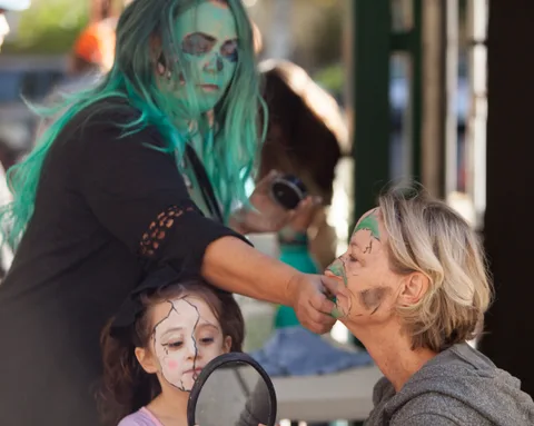 A woman with green-tinted hair applies zombie face paint to an older woman under a market awning.