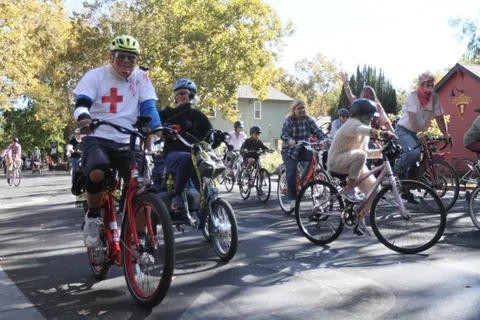 A rider with a red cross armband pedals with the group past a red building.
