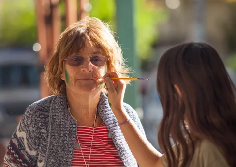 A woman with glasses has face paint brushed onto her cheek.
