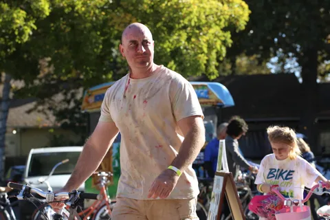 A bald man with green paint on his shirt and a festival wristband walks smiling toward the camera.