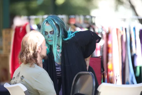 A woman with teal hair and zombie face paint leans down to talk with a seated man.