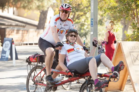 Two riders in matching orange jerseys sit on a tandem bike as a costumed figure passes behind.