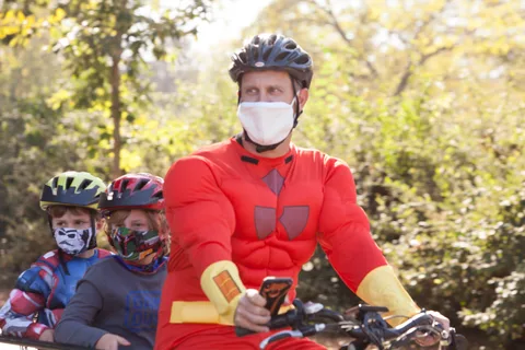 A cyclist in a red superhero costume rides past a row of masked kids on bikes.