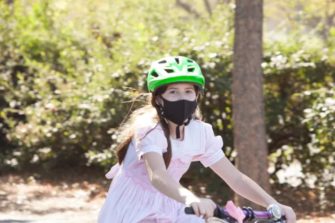 A girl in a pink dress and green helmet rides her bike straight toward the camera.