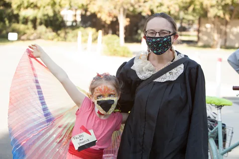 A girl with a painted mask stands next to a woman in a black robe costume as colorful fabric streams overhead.