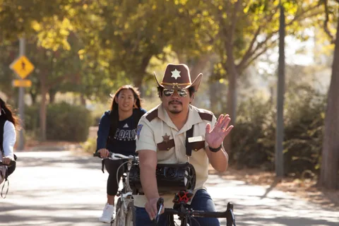 A man in a cowboy hat and sunglasses waves while riding his bike.