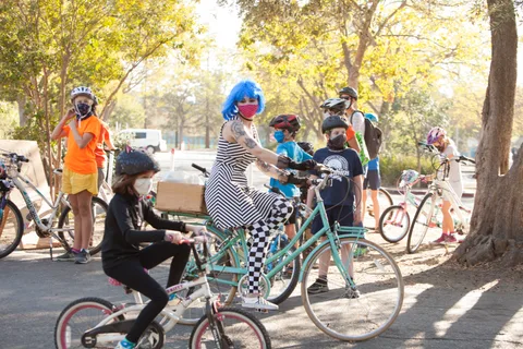 Kids in striped and checkered costumes ride bikes together on the park path.