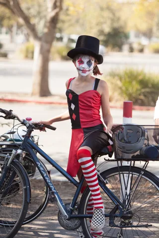 A rider in a red and black harlequin costume and top hat with painted clown makeup sits on her bike.