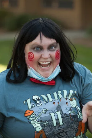 A woman with red spiral cheek paint grins widely, mask pulled below her chin, wearing an official Zombie Bike Parade shirt.