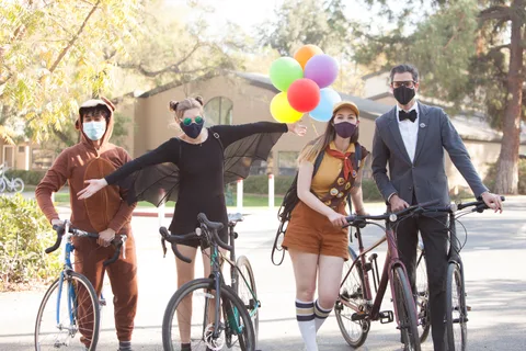 Four costumed cyclists with balloons and matching bow tie pose together in golden light.