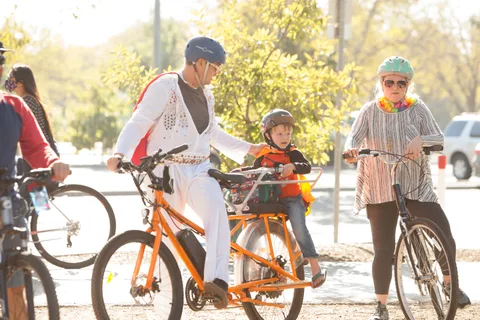 A man in an Elvis costume rides an orange tandem cargo bike with a child passenger.
