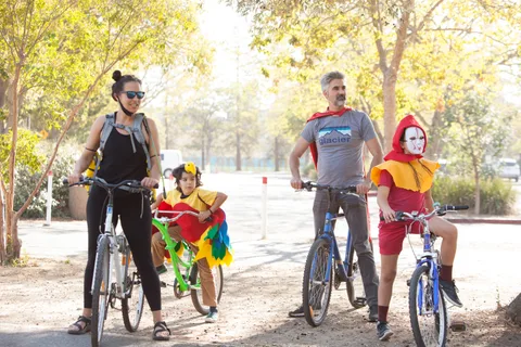 A child in a colorful parrot costume sits on a bike between two adults.