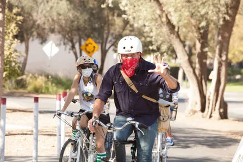 A man in a red bandana mask waves with kids and riders nearby.