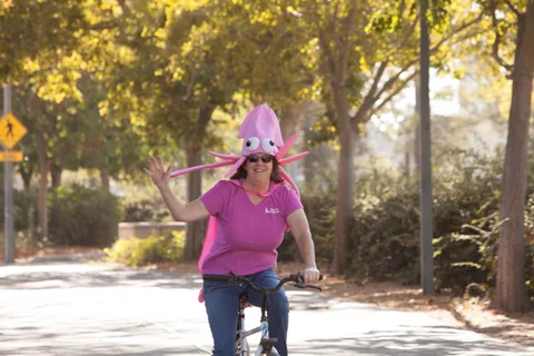 A woman in a pink octopus hat waves while riding her bike.
