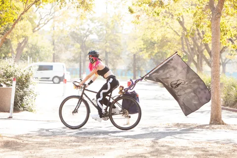 A masked rider in black and white streetwear pedals hard while a large flag streams behind the bike in golden light.