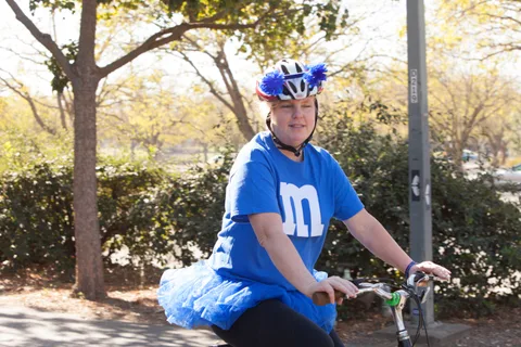 A kid in a puffball helmet cover and blue tutu rides a bike.