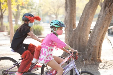 A child in a pink costume rides ahead while an adult in a red devil costume follows on foot.