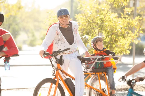 A man in a white Elvis-style jumpsuit rides a tandem bike with a child.