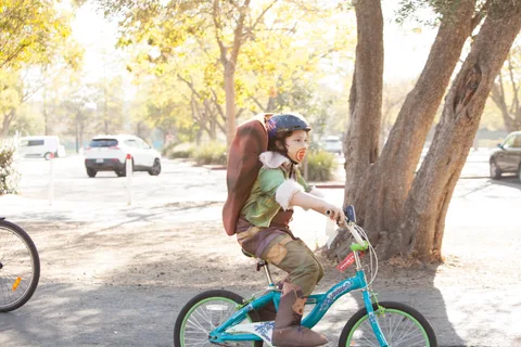 A child rides a teal bike away down a sunlit tree-lined path.