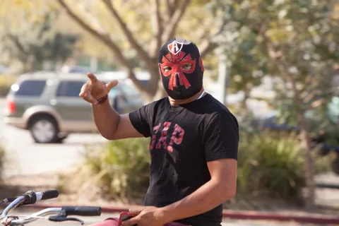 A man in a red wrestling mask gestures while standing with his bike.