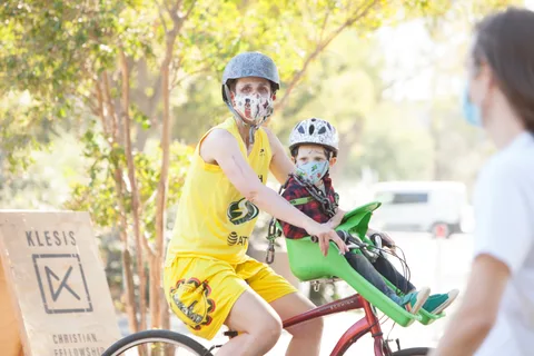 An adult in yellow rides with a child on a trailer bike as a passerby crosses in front.