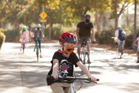 A boy in a red helmet and orange mask rides while another cyclist passes behind.