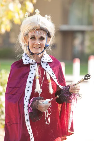 A woman in a jeweled crown and ermine-trimmed royal cape smiles warmly at the camera.