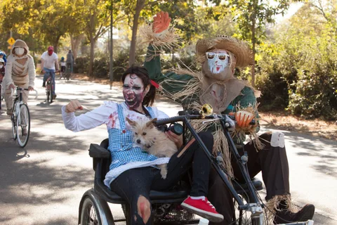 A zombie Dorothy waves from a tandem trike carrying a small dog while a scarecrow pedals behind her.