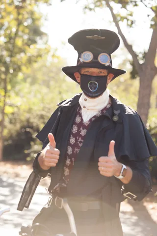 A man in a black top hat and steampunk goggles wears a bandana mask over his face.