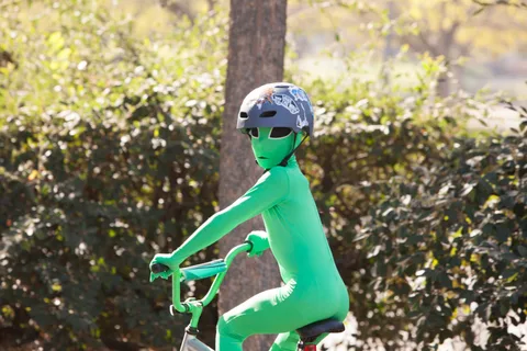A child in a full green alien bodysuit rides a small bike.