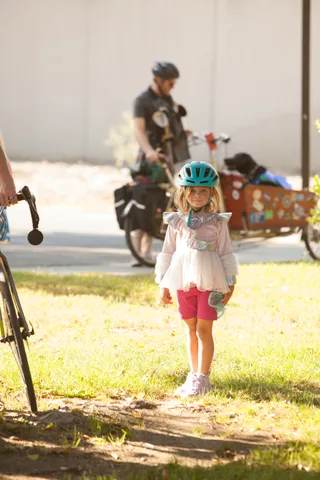 A girl in a pink fairy costume walks past a rider towing a cargo trailer.
