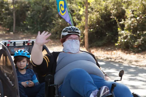 A woman on a recumbent trike waves while a child rides behind in a covered trailer.
