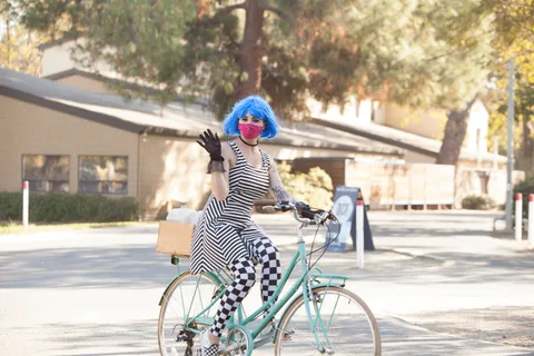 A woman in a blue wig and black and white checkered leggings waves while riding a teal bike.