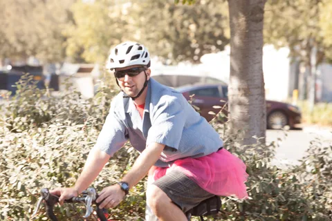 A man in a pink tutu over shorts rides a bike.