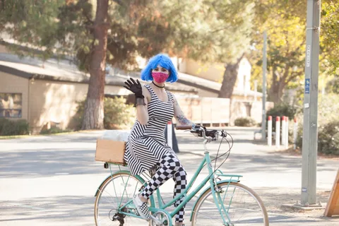 A woman in a blue wig and checkered leggings waves from a teal cruiser bike.