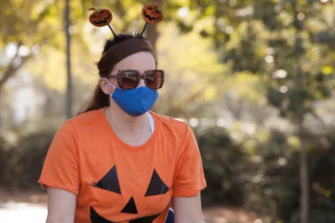 A woman in a pumpkin-print shirt and jack-o-lantern headband wears a blue mask.