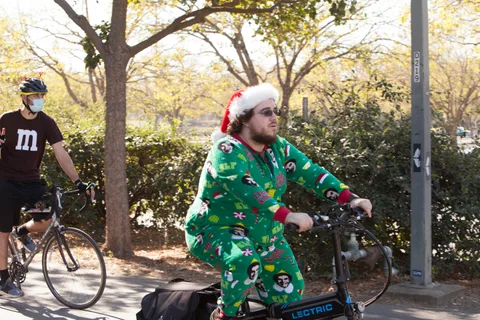 A man in a green holiday-print onesie and Santa hat rides a bike.