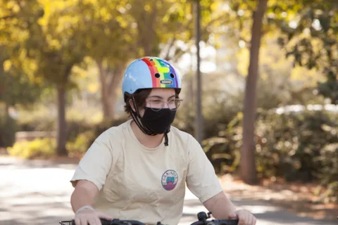 A kid in a rainbow-striped helmet cover and mask rides a bike.