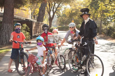 A rider in a dark robed costume greets a group of masked kids with their bikes.