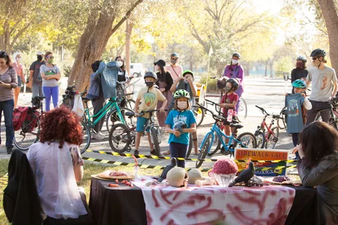 A crowd of masked riders waits behind caution tape near a table decorated with fake skulls and brains.
