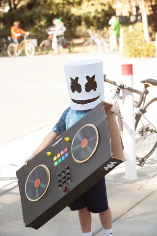 A kid wears a full Marshmello head costume and carries a handmade cardboard DJ deck.