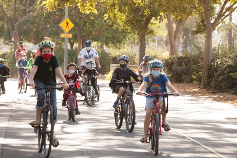 A line of masked kids and adults ride bikes toward the camera through golden light past a crosswalk sign.