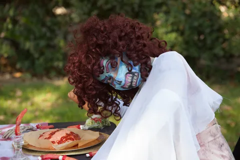 A woman with a curly red wig and a painted blue skull face leans over a table set with fake brains and severed fingers.
